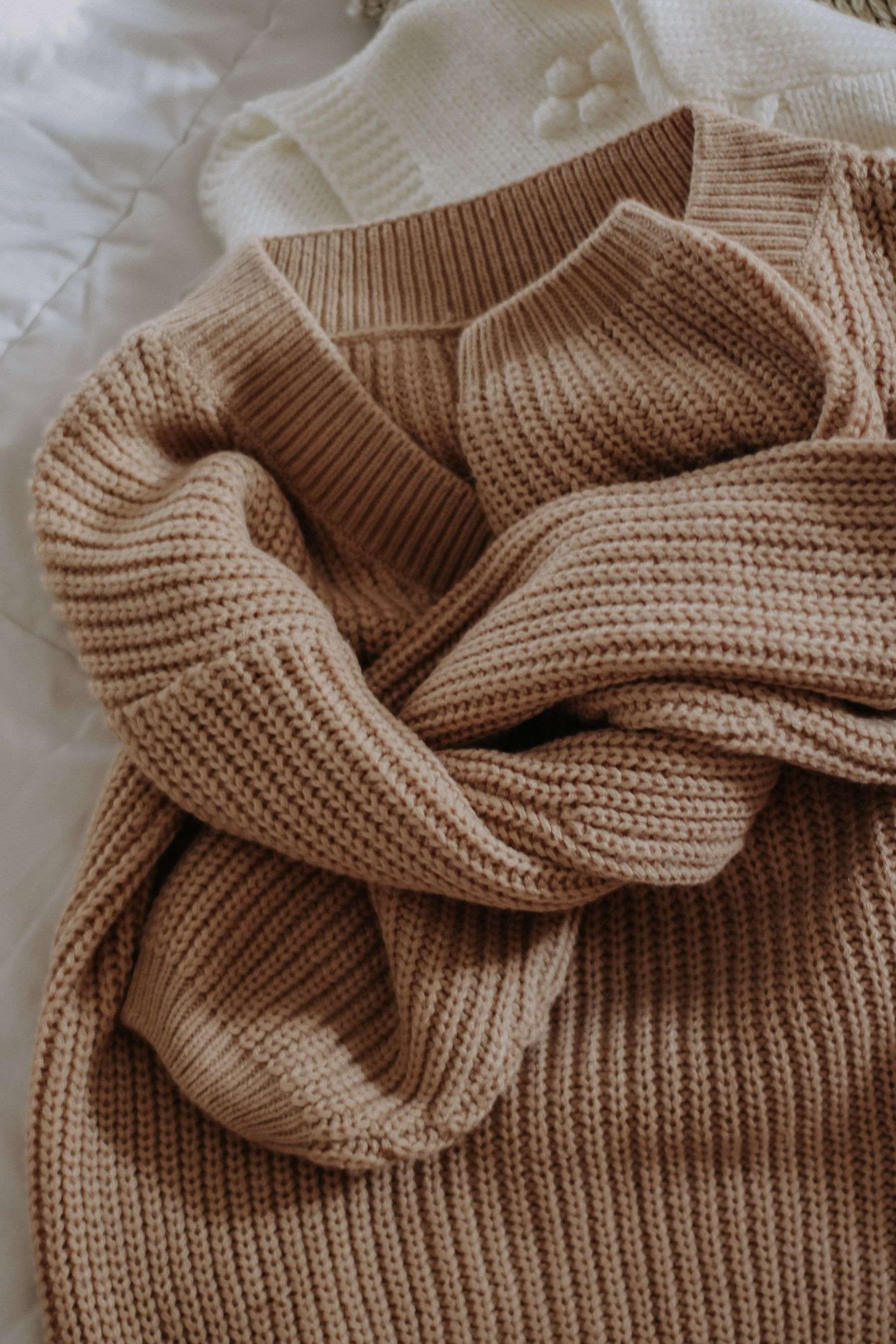 A camel rib-knit sweater loosely folded on a white bed