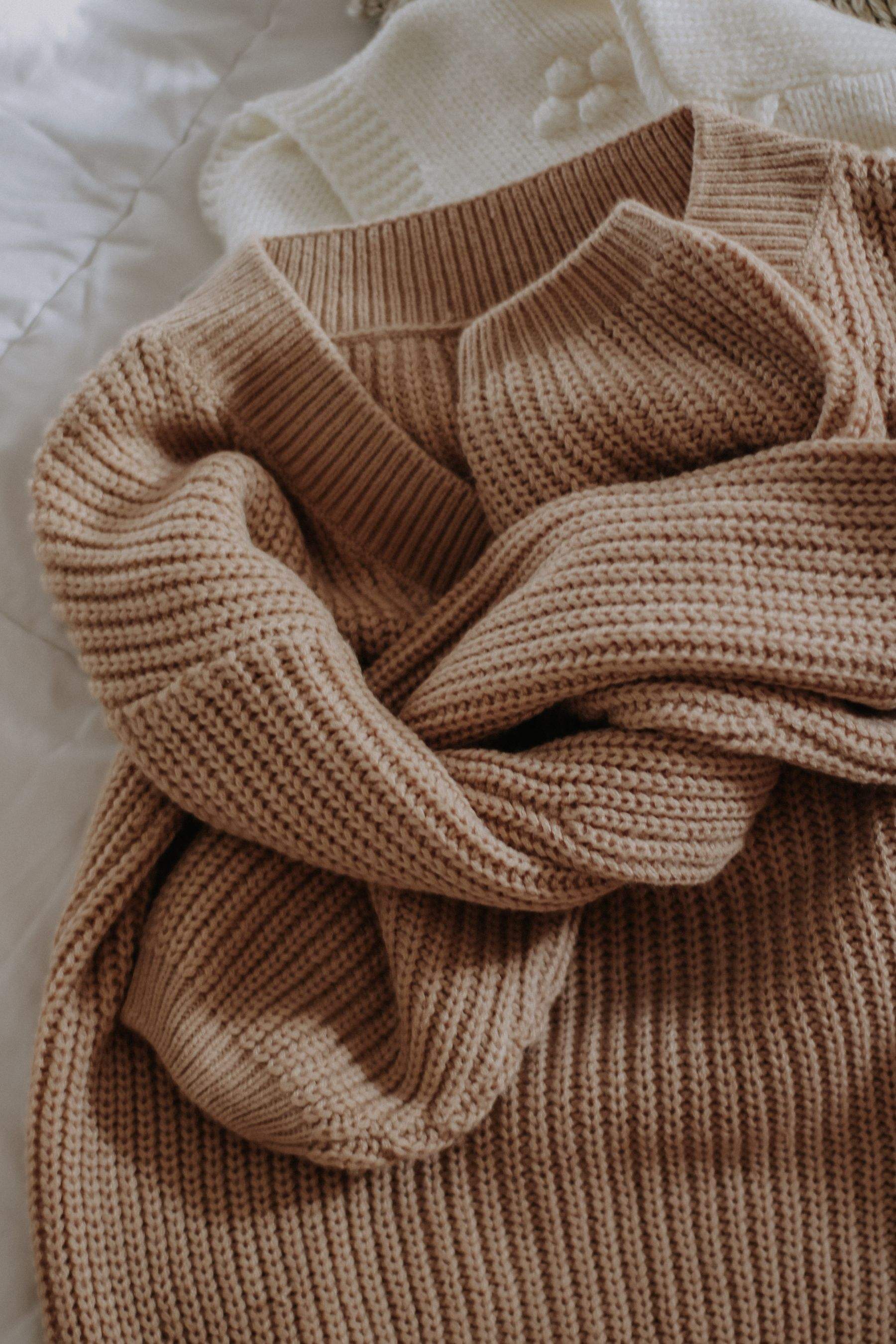 A camel rib-knit sweater loosely folded on a white bed
