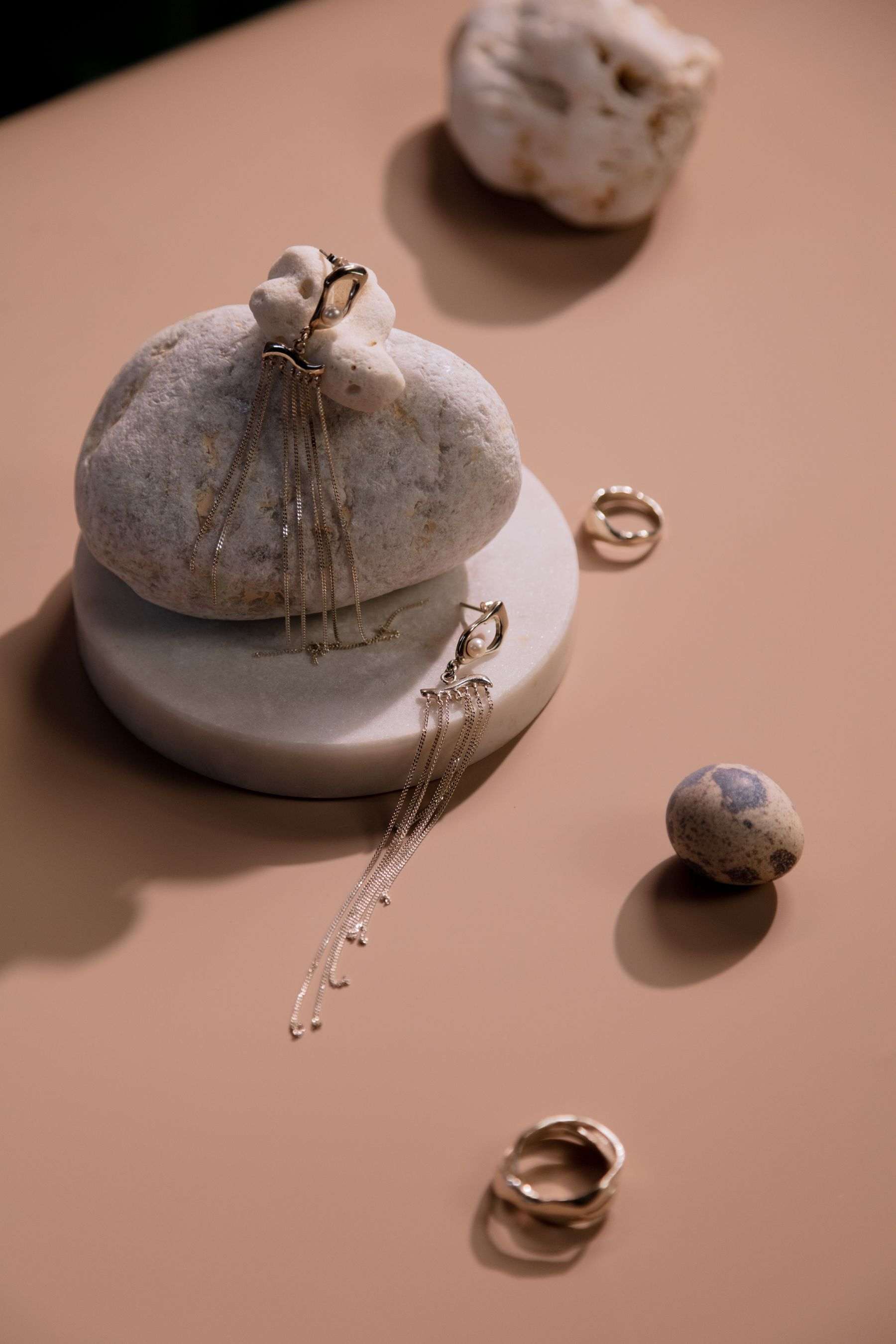 Fine chain drop earrings draped over a pale pebble on a stone plinth, with small rings on a peach surface