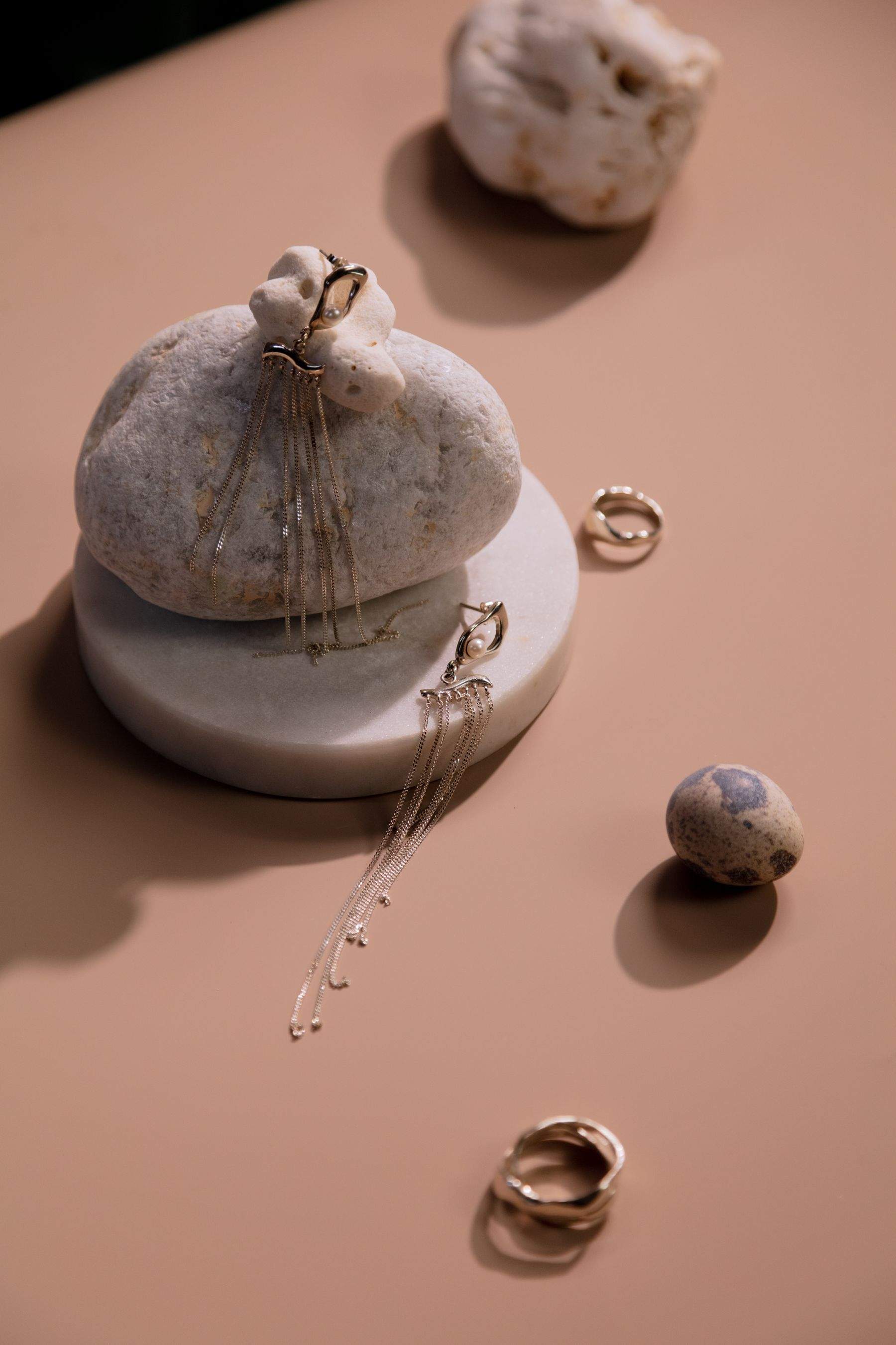 Fine chain drop earrings draped over a pale pebble on a stone plinth, with small rings on a peach surface