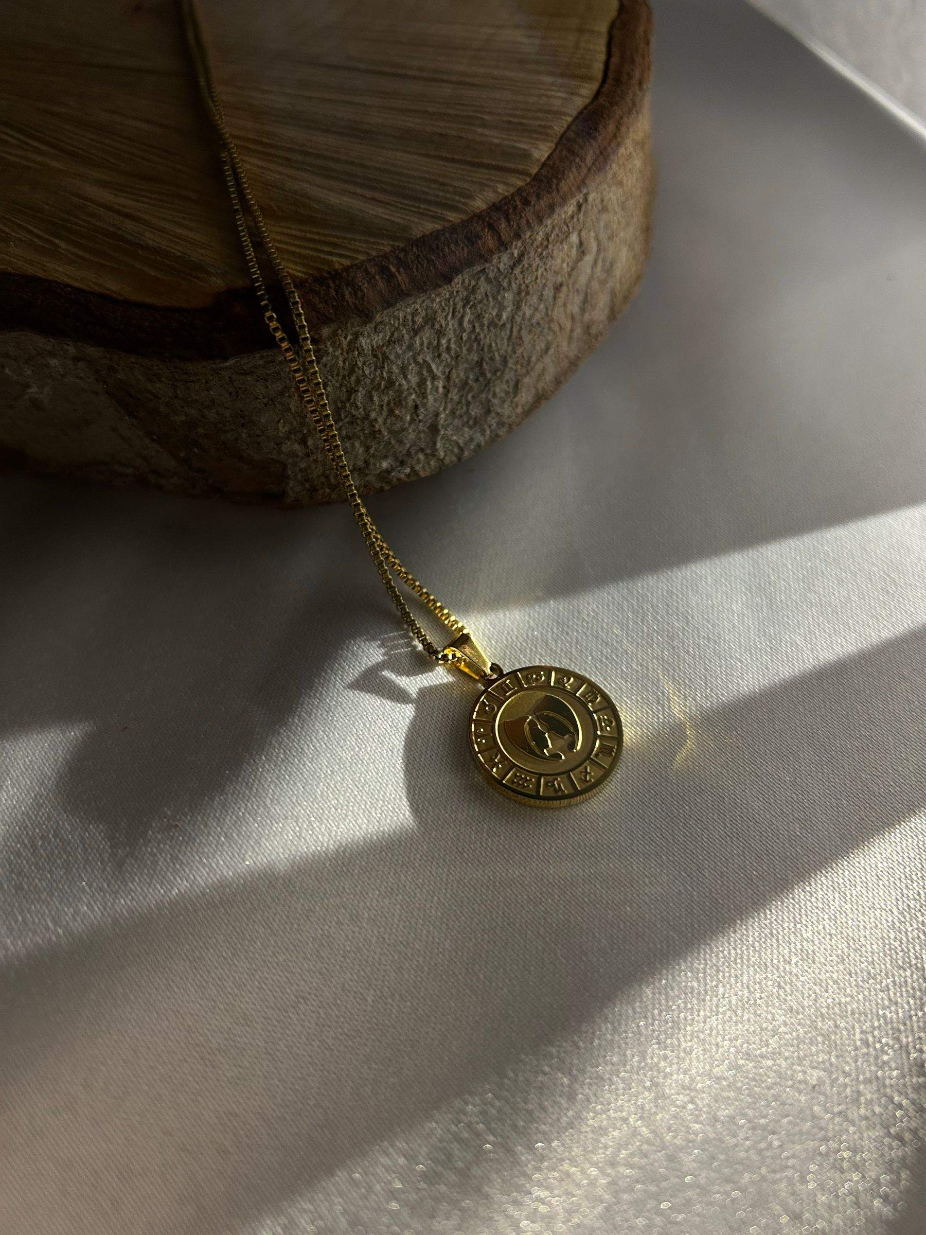 A gold coin pendant on a fine chain, lying in soft light and shadow beside a slice of wood