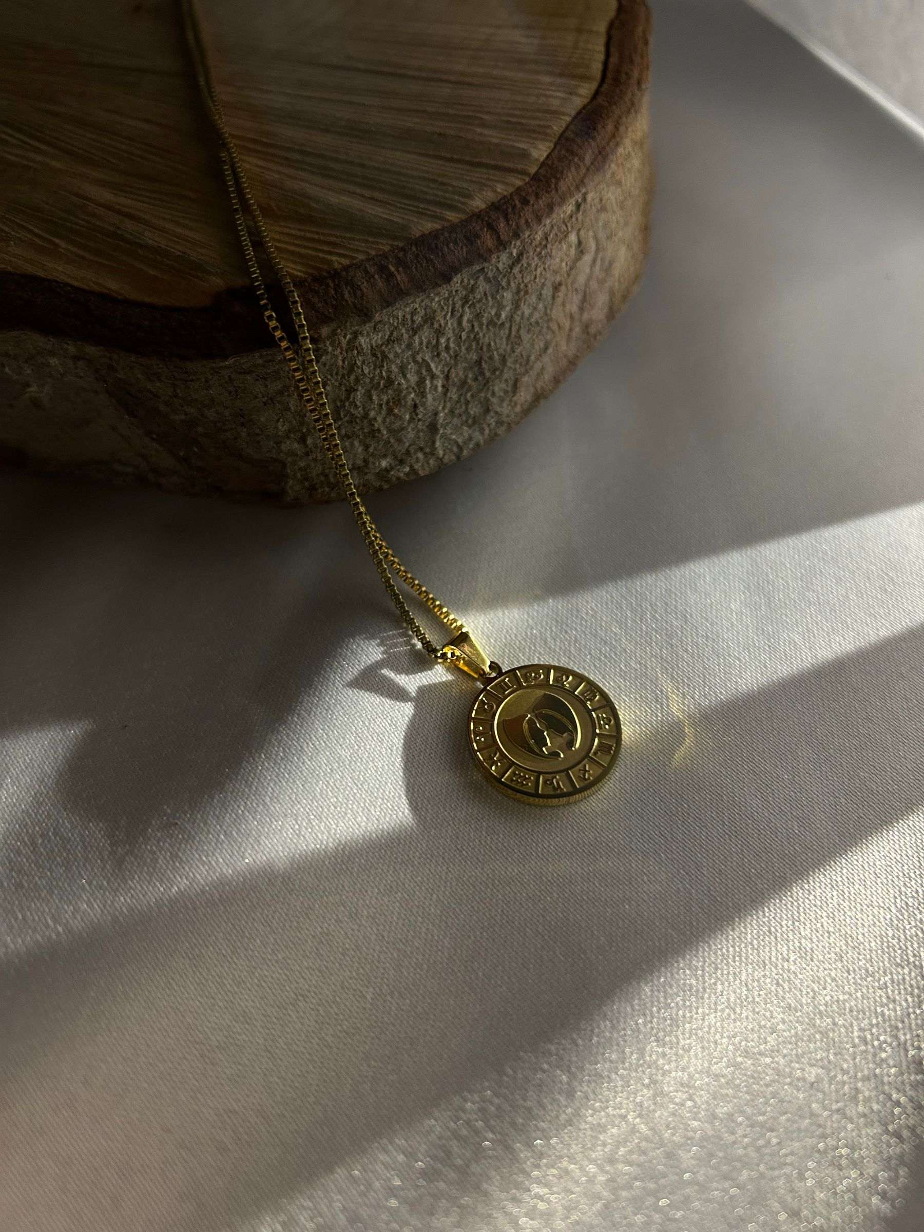 A gold coin pendant on a fine chain, lying in soft light and shadow beside a slice of wood