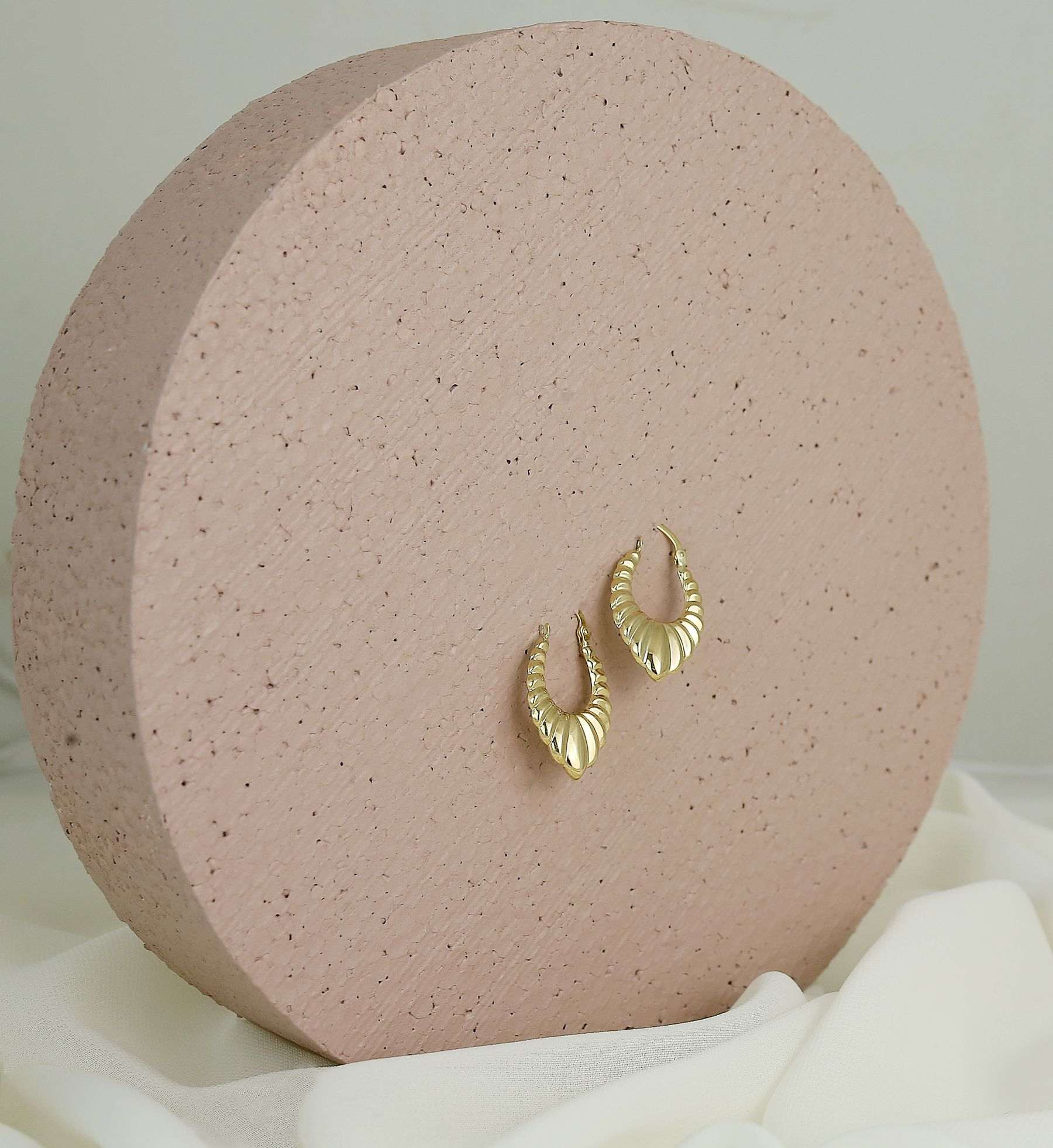 A pair of ribbed gold crescent hoops resting on a blush speckled stone disc