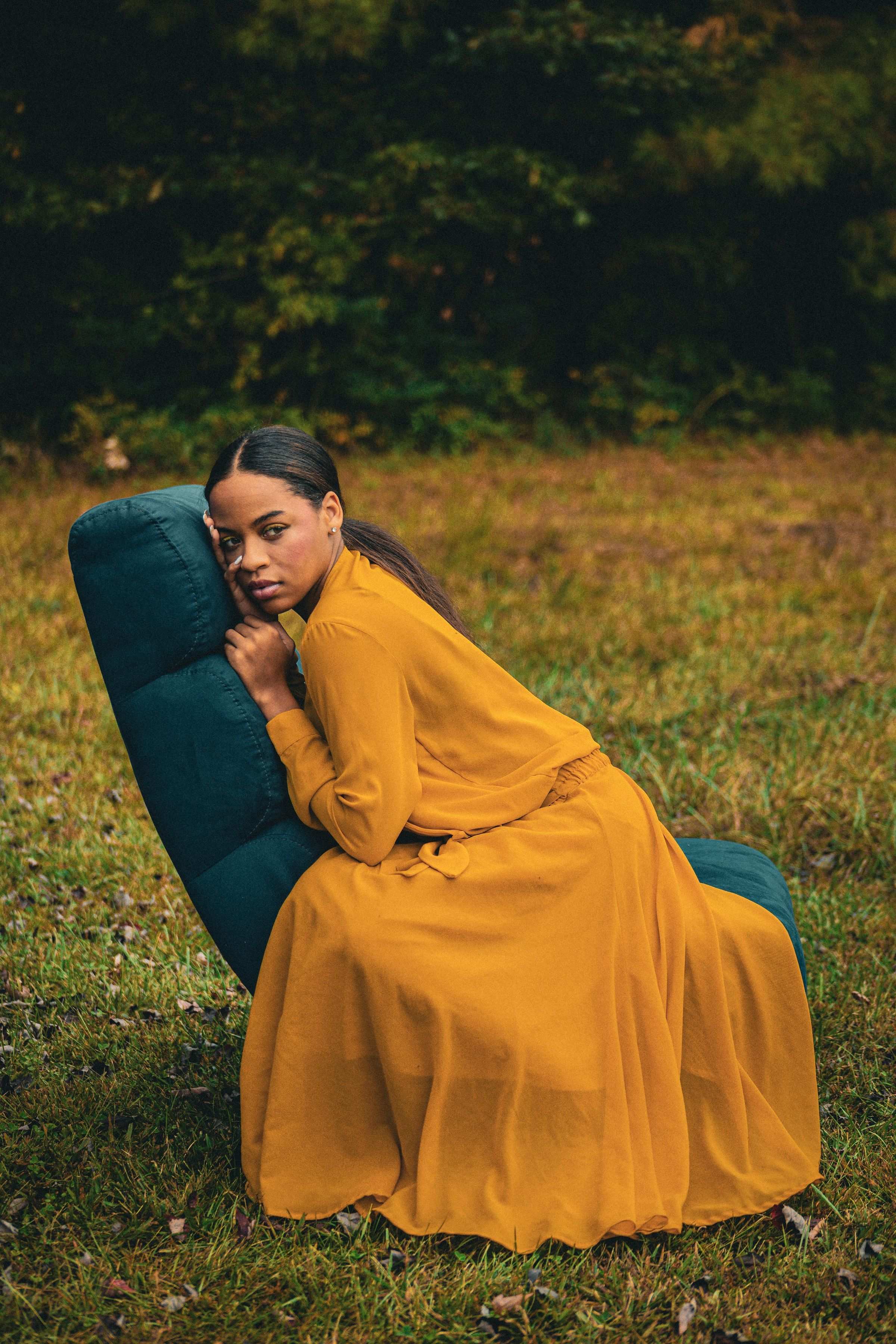 A woman in a flowing marigold dress resting against a moss green armchair in a meadow