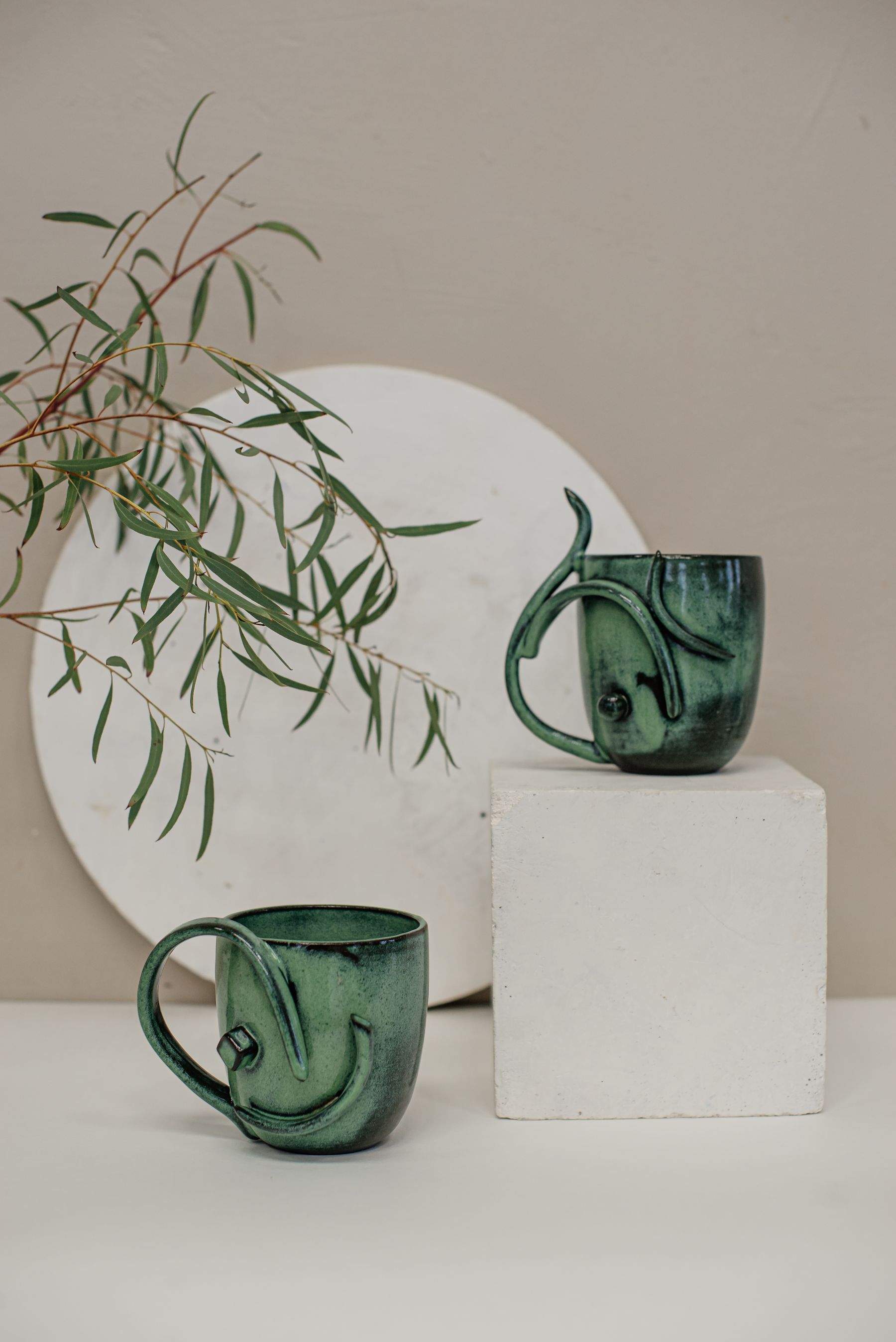 Two hand-glazed moss green mugs beside a eucalyptus branch