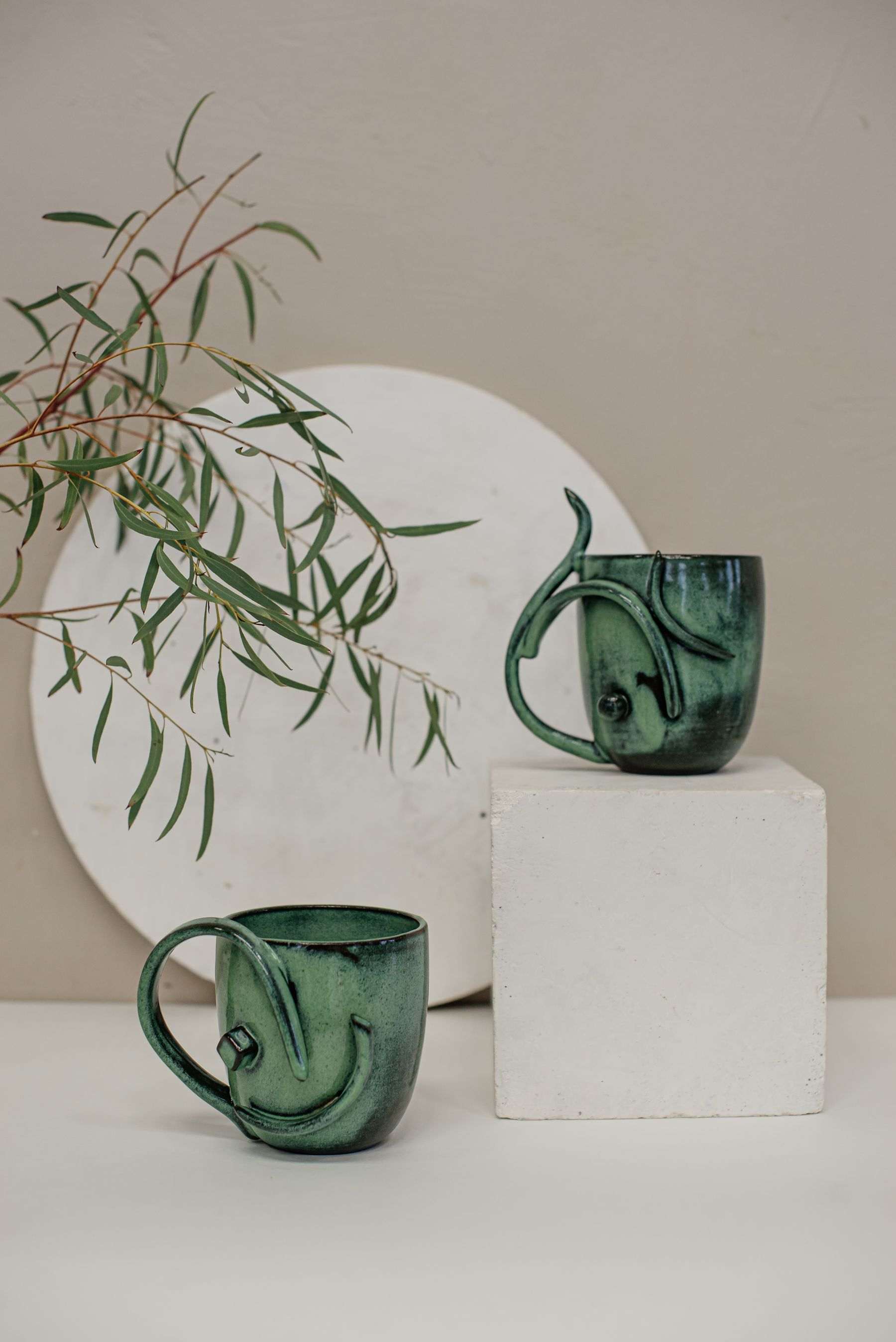 Two hand-glazed moss green mugs beside a eucalyptus branch
