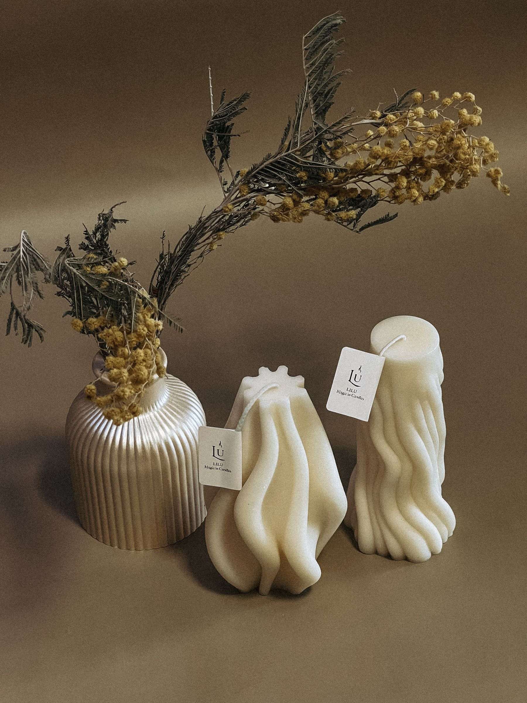 Two cream sculptural pillar candles, one twisted and one wave-shaped, beside dried golden flowers