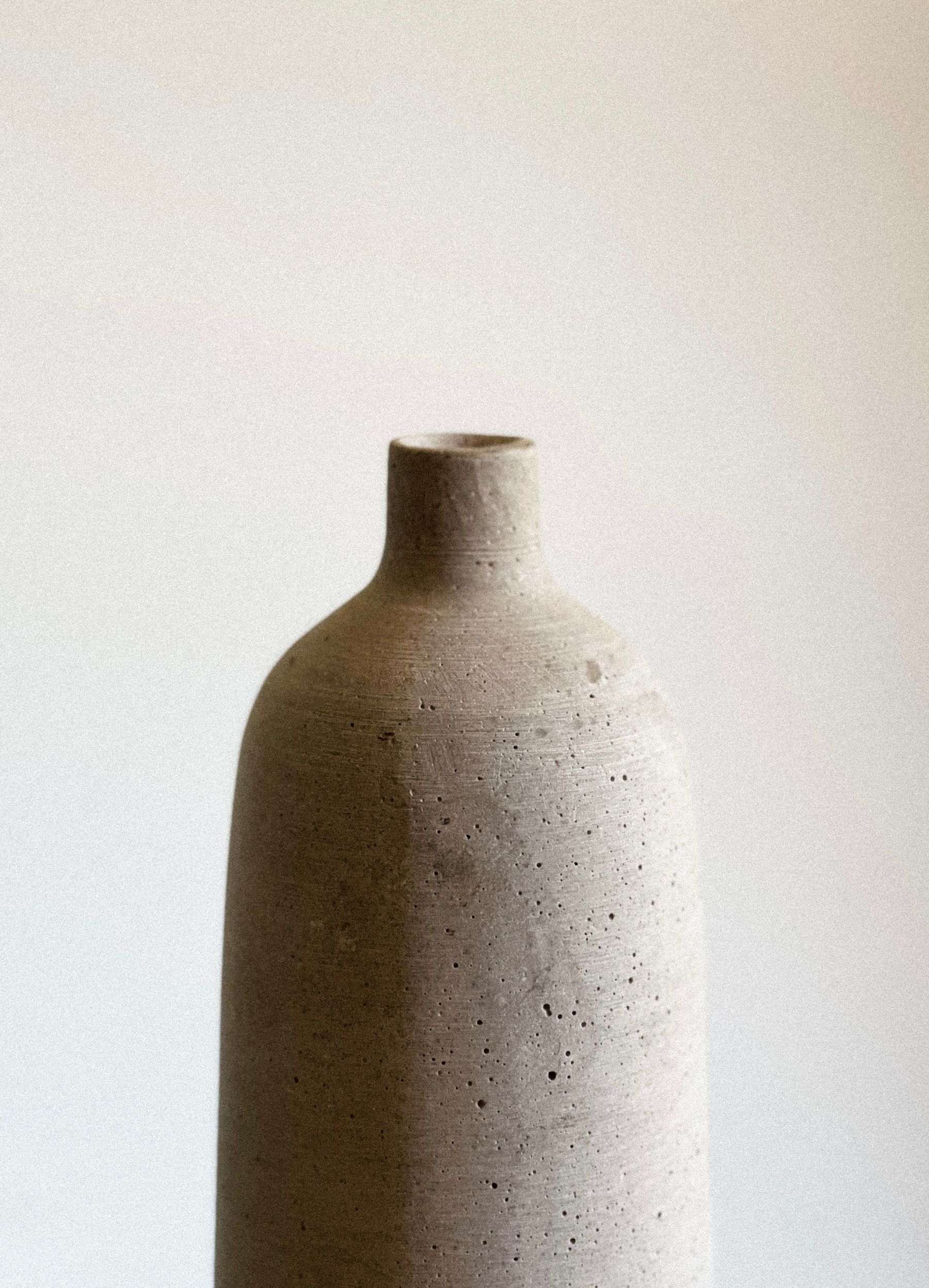 A tall, unglazed speckled stoneware bottle vase against a white wall