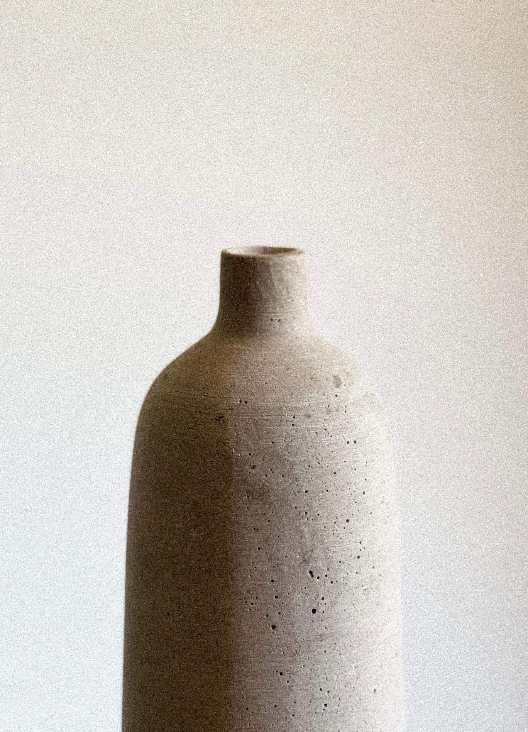 A tall, unglazed speckled stoneware bottle vase against a white wall