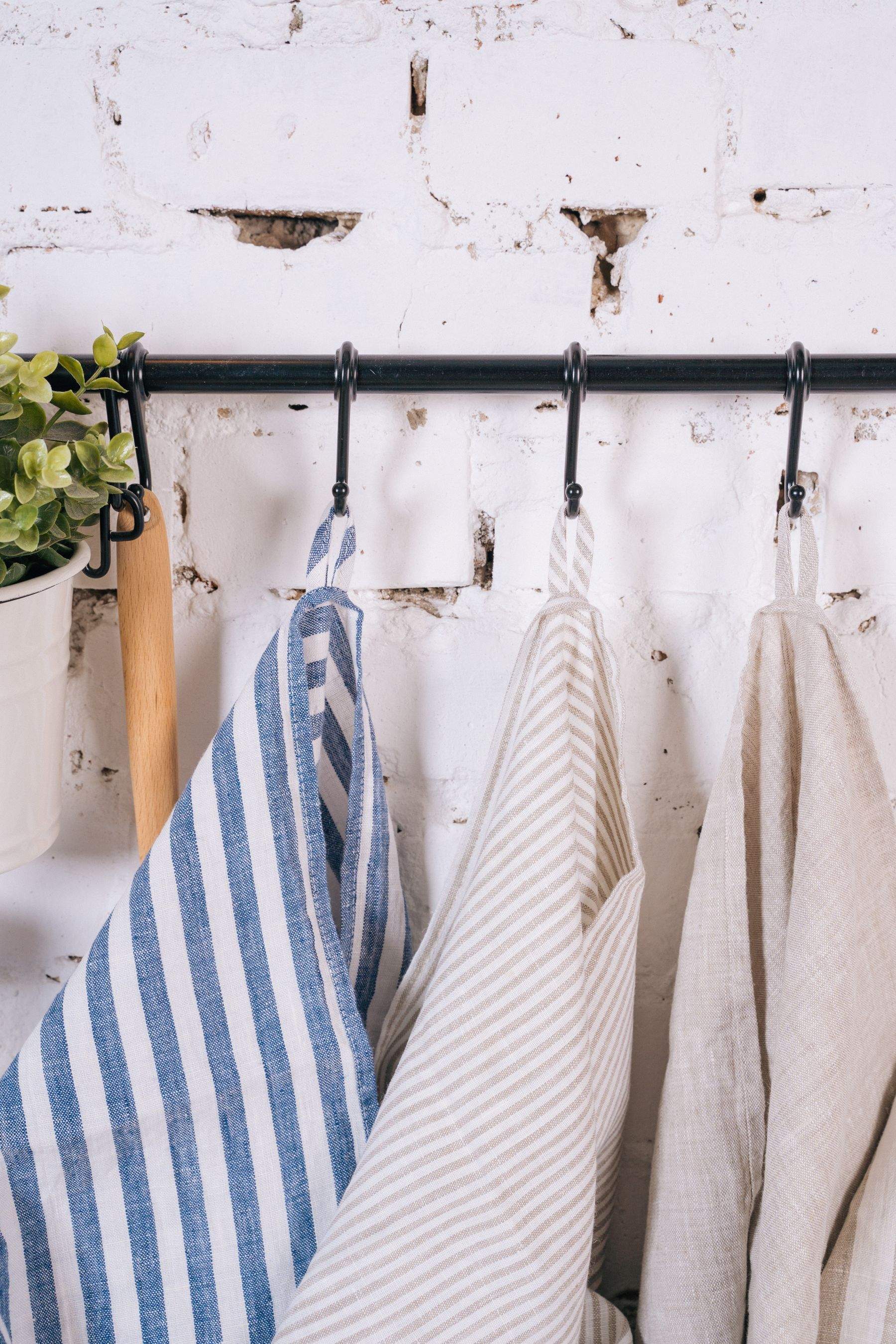 Blue striped, ticking-stripe and plain natural linen tea towels hanging from hooks on a whitewashed brick wall