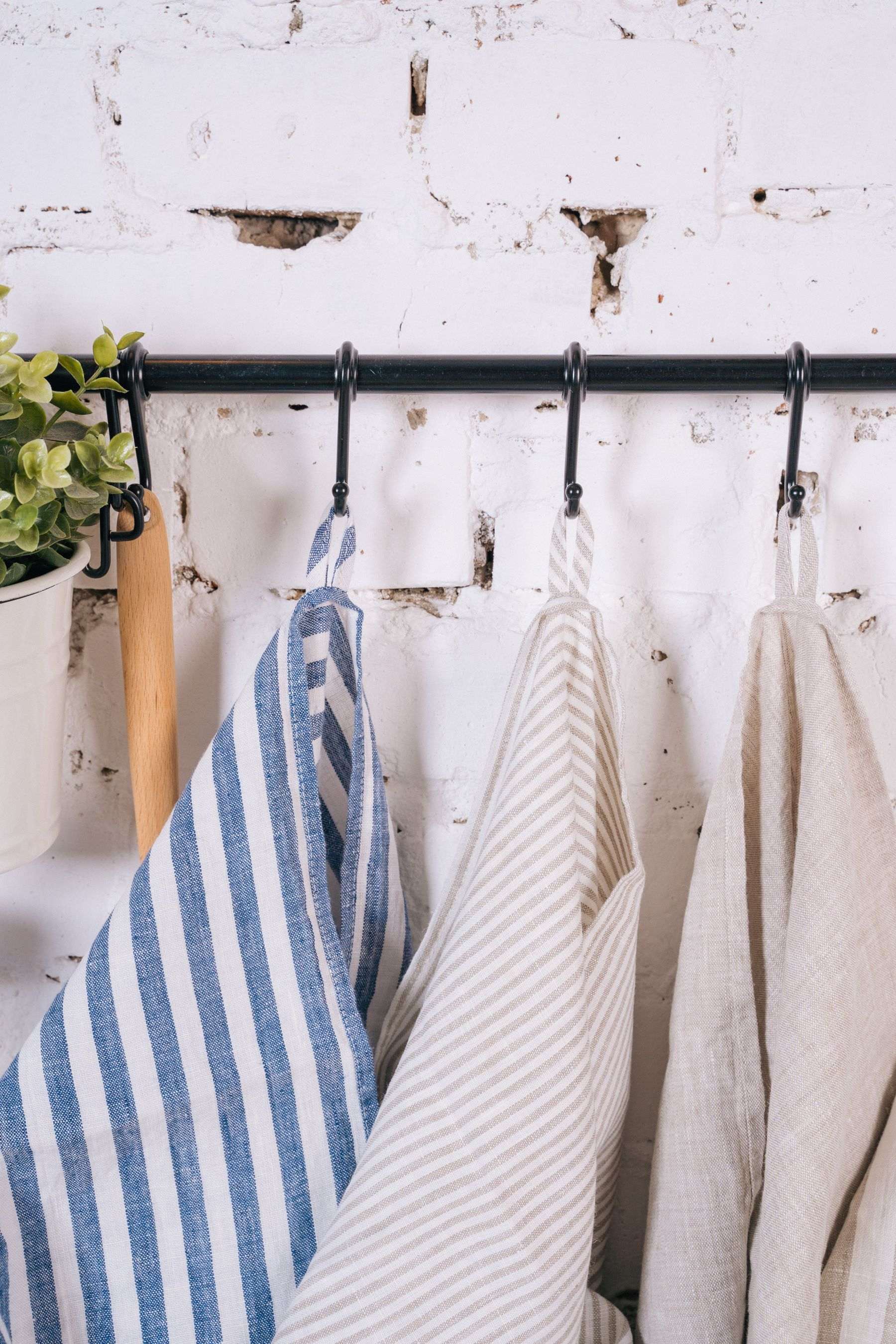 Blue striped, ticking-stripe and plain natural linen tea towels hanging from hooks on a whitewashed brick wall